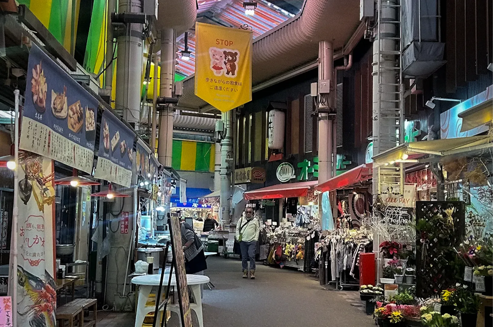 Covered shopping arcade in Kanazawa, Japan, featuring seafood stalls, flower shops, local vendors, and pedestrians walking beneath a colorful translucent roof in a traditional market district.