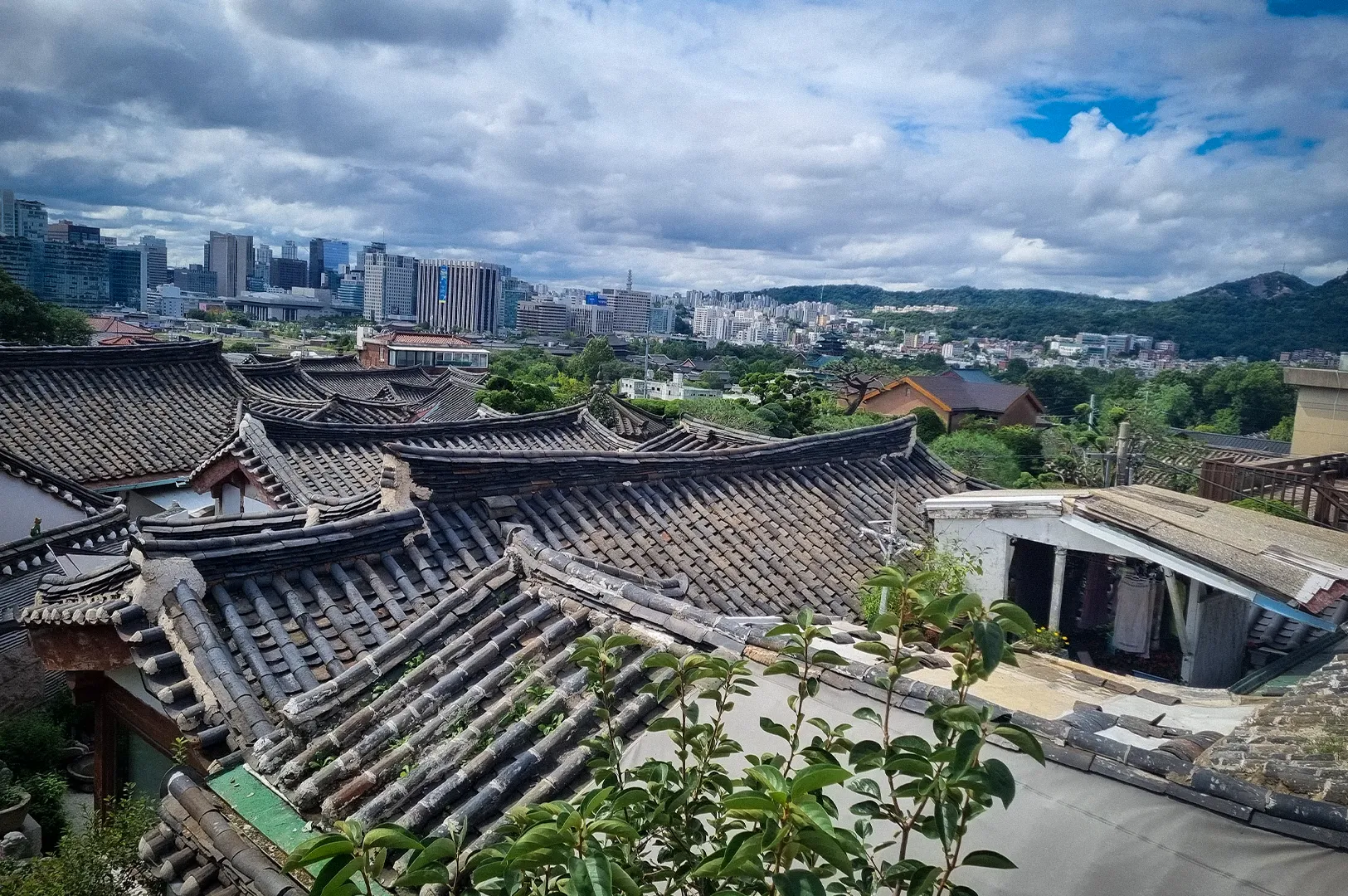 Panoramic view of traditional Korean hanok rooftops in Bukchon Hanok Village with modern Seoul skyscrapers and green hills in the background under a dramatic cloudy sky.