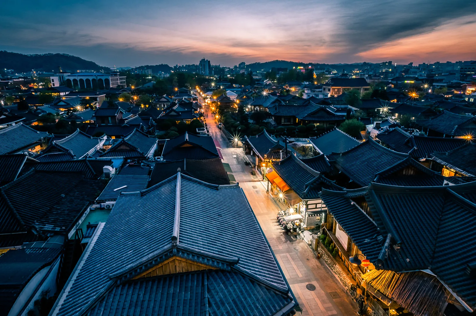 Aerial night view of Jeonju Hanok Village in South Korea featuring illuminated traditional hanok rooftops, vibrant streets and a colorful sunset skyline.