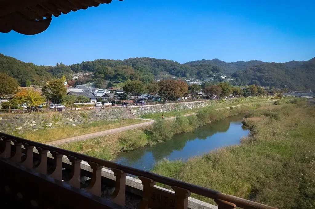 Scenic view of Jeonju Hanok Village from a traditional pavilion, overlooking a tranquil river, walking paths, historic hanok rooftops, and forested hills under a clear blue sky.