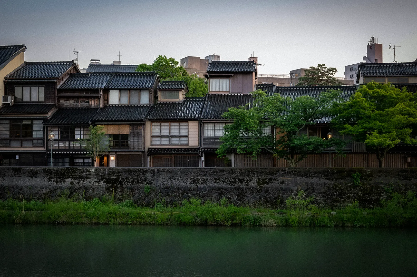 Traditional Japanese houses along a riverside in Kanazawa, Japan, featuring historic architecture, tiled roofs, lush greenery, and a peaceful waterfront setting.