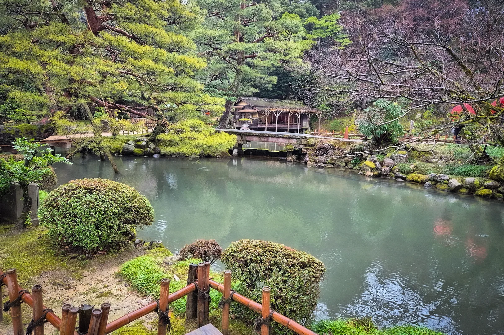 Scenic pond surrounded by manicured shrubs, pine trees, moss-covered rocks, and a traditional wooden pavilion in Kenrokuen Garden, Kanazawa, Japan.