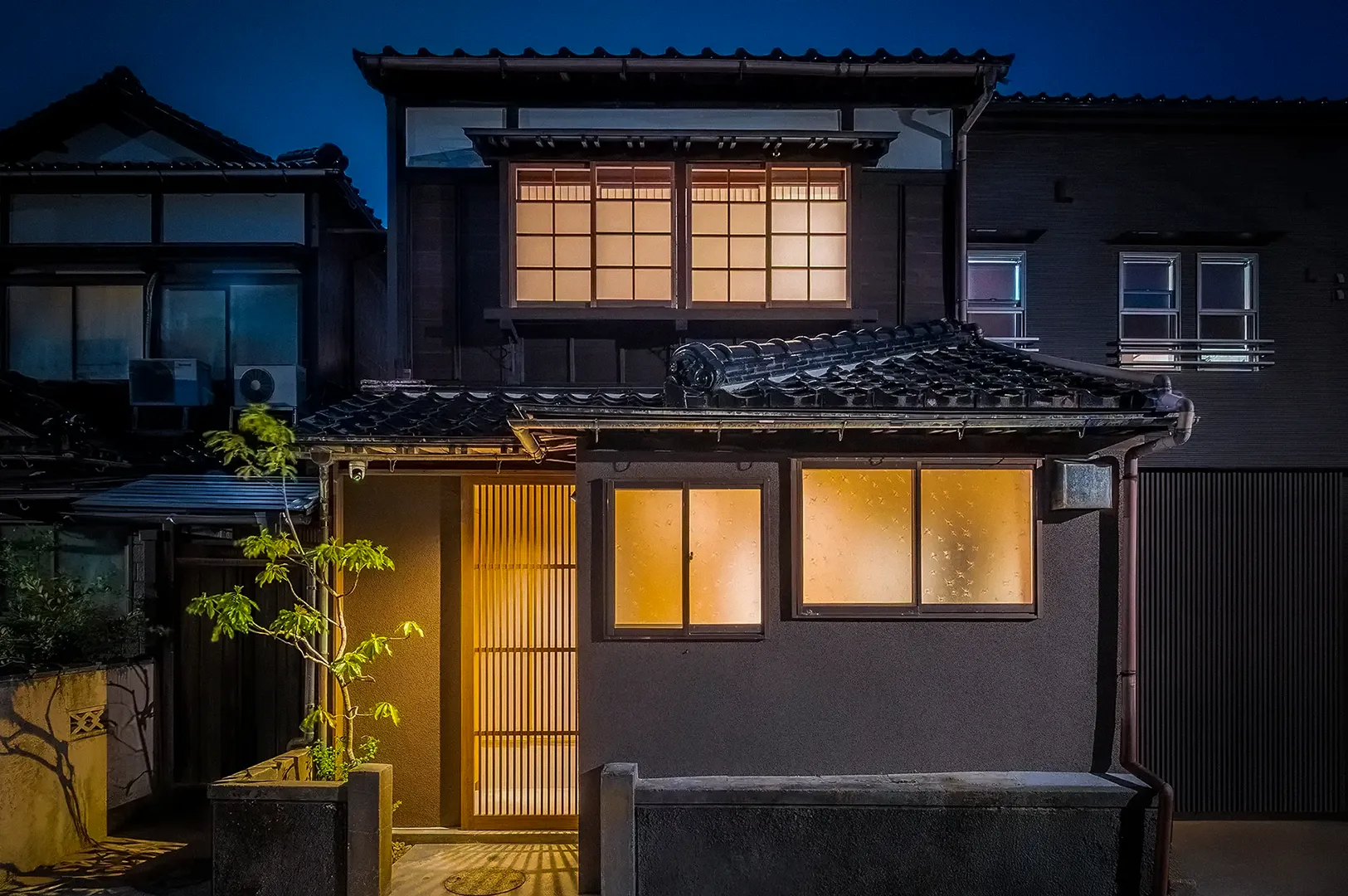 Traditional Japanese machiya townhouse in Kanazawa illuminated at night, featuring wooden lattice doors, shoji-style windows, tiled roofing, and warm interior lighting characteristic of a renovated heritage residence.