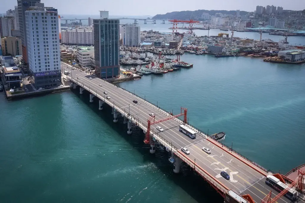 Aerial view of Yeongdo Bridge connecting Yeongdo Island to downtown Busan, with the harbor, fishing boats, waterfront skyline, and coastal cityscape in South Korea.