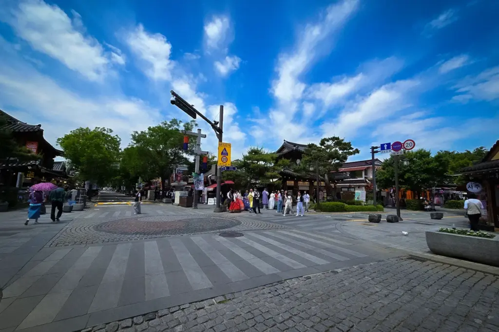 Wide view of Jeonju Hanok Village main street with traditional Korean architecture, visitors in hanbok, cobblestone roads, and dramatic blue skies with scattered clouds.