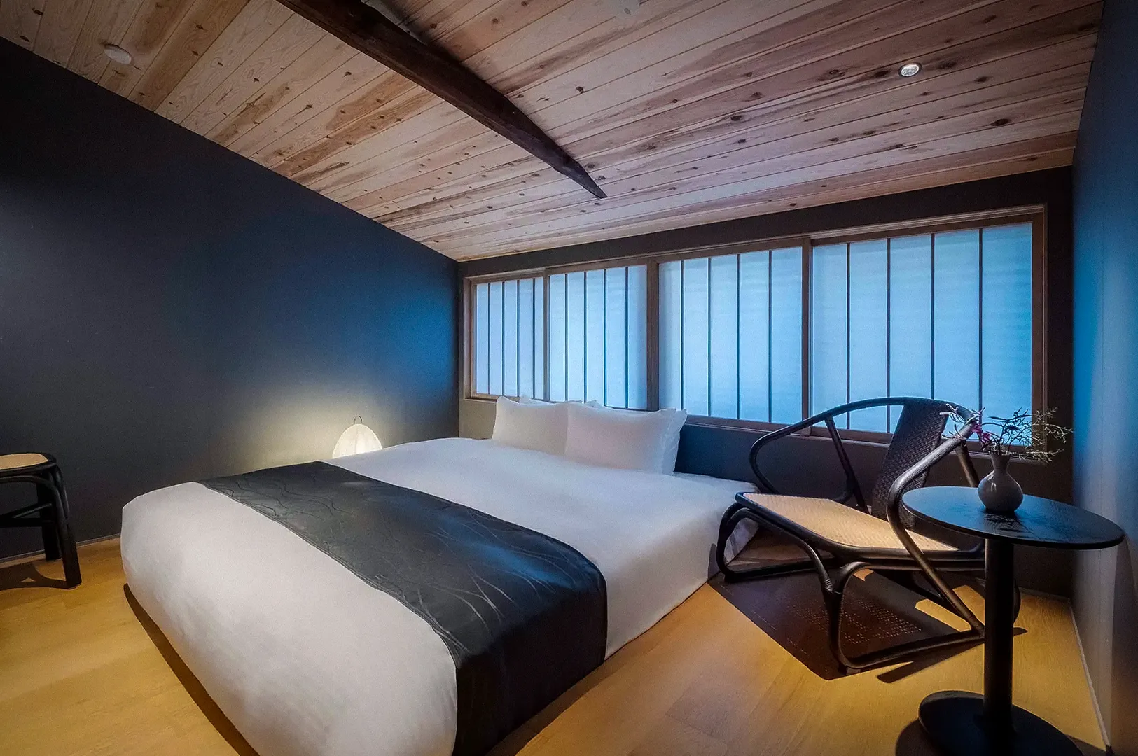 Modern bedroom inside a traditional machiya townhouse in Kanazawa, Japan, featuring a large bed, shoji-style windows, wooden ceiling beams, minimalist furnishings, and warm contemporary Japanese design.