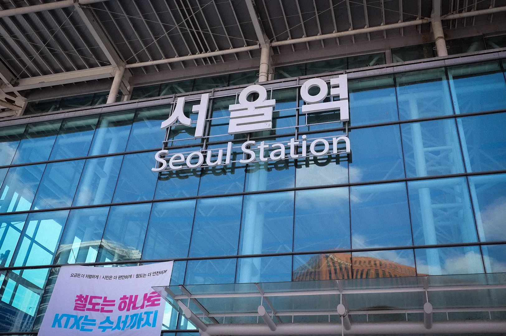 Exterior facade of Seoul Station featuring large Korean and English signage on a modern glass building, a major transportation hub connecting KTX high-speed rail, subway, and regional train services in Seoul, South Korea.