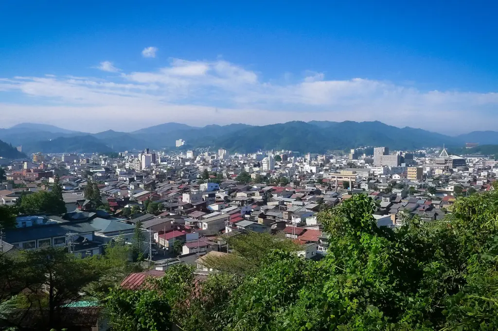 Panoramic view of Takayama, Japan, showcasing the city's rooftops, surrounding forested mountains, and clear blue skies from an elevated viewpoint.