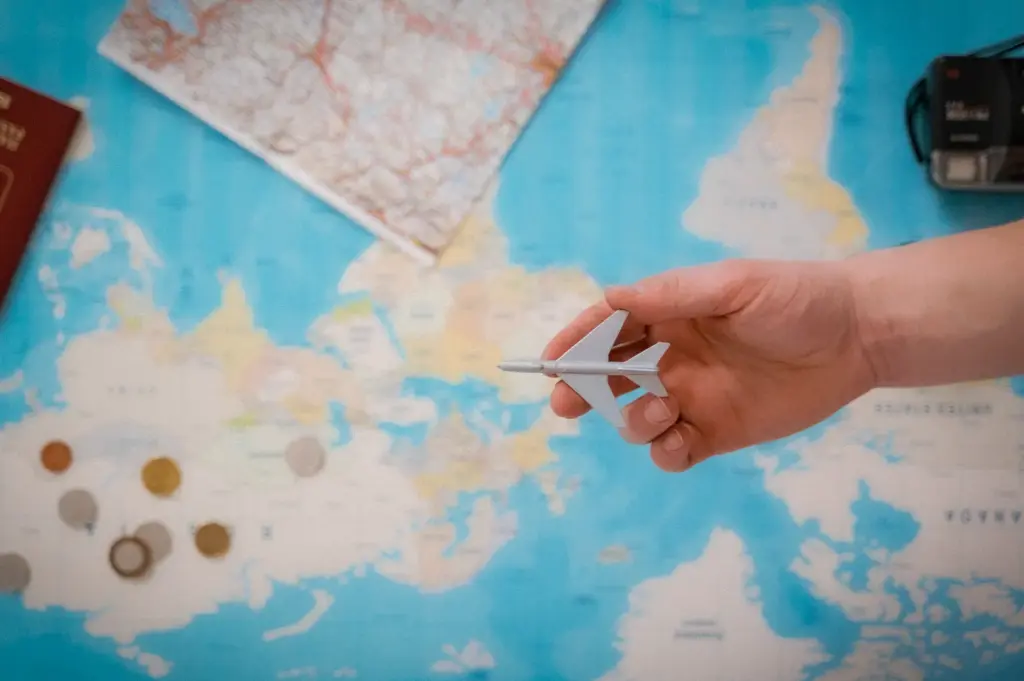 Hand holding a model airplane over a world map with a passport, travel map, camera, and foreign coins, symbolizing international travel planning and vacation preparation.