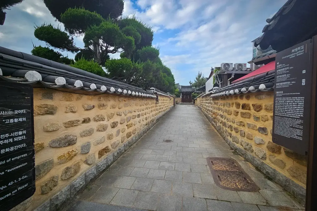 Traditional stone wall alley in Jeonju Hanok Village featuring tiled roofs, manicured pine trees, historic Korean architecture, and a peaceful pedestrian walkway under a cloudy sky.