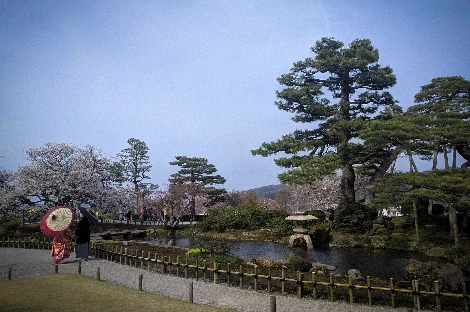 Kenroku-en Garden in Kanazawa, Japan, featuring a traditional landscaped pond, stone lantern, pine trees, cherry blossoms, and visitors in traditional Japanese attire.
