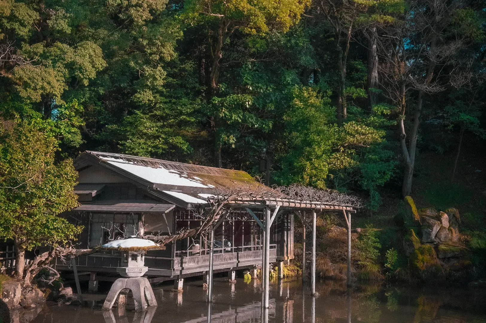 Traditional Japanese teahouse overlooking a tranquil pond in Kenroku-en Garden, Kanazawa, Japan, surrounded by lush trees, stone lanterns, and reflections on the water.