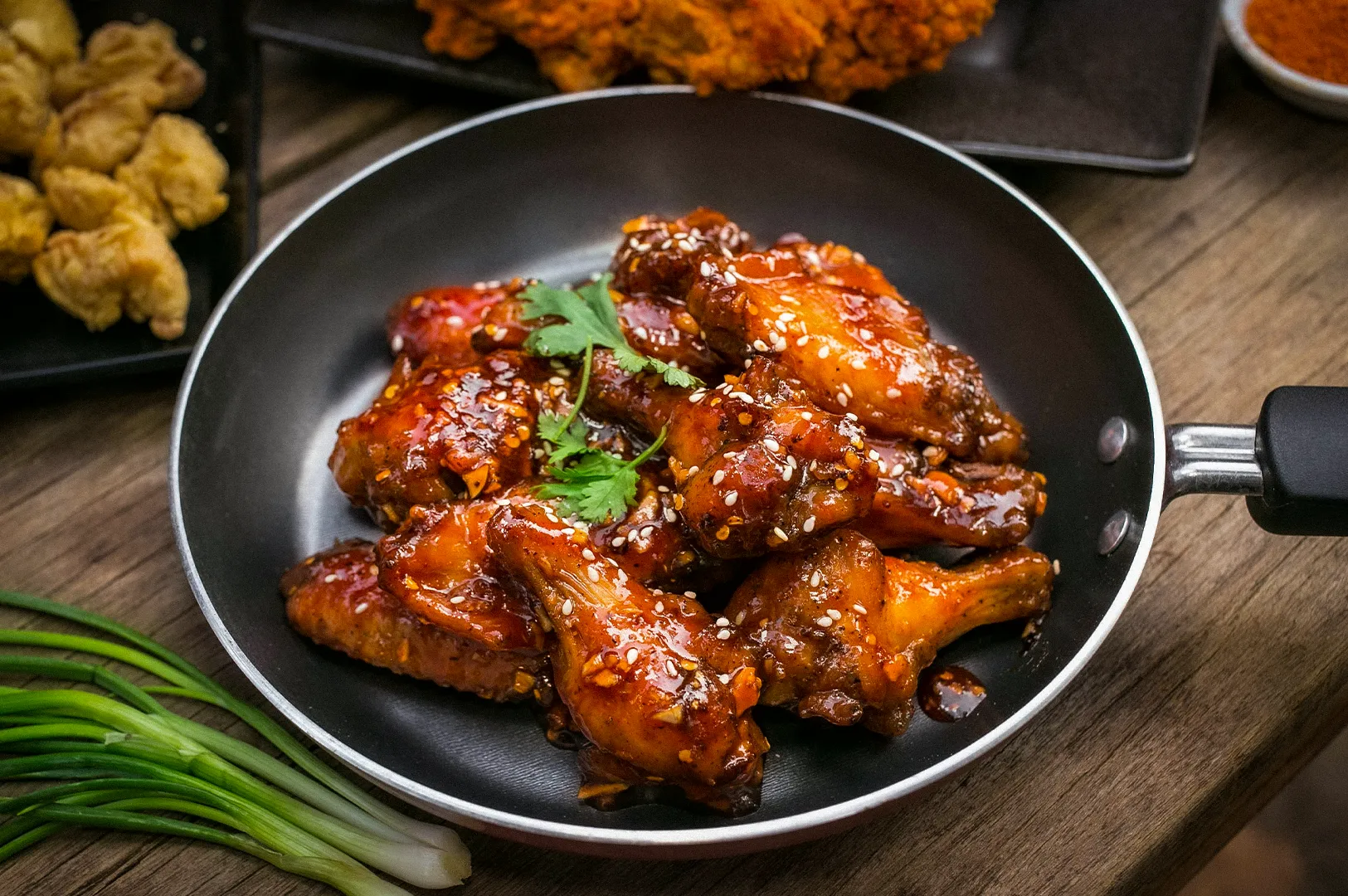 Crispy Korean fried chicken wings coated in a glossy sweet and spicy sauce, garnished with sesame seeds and fresh herbs, served in a skillet with side dishes on a rustic wooden table.