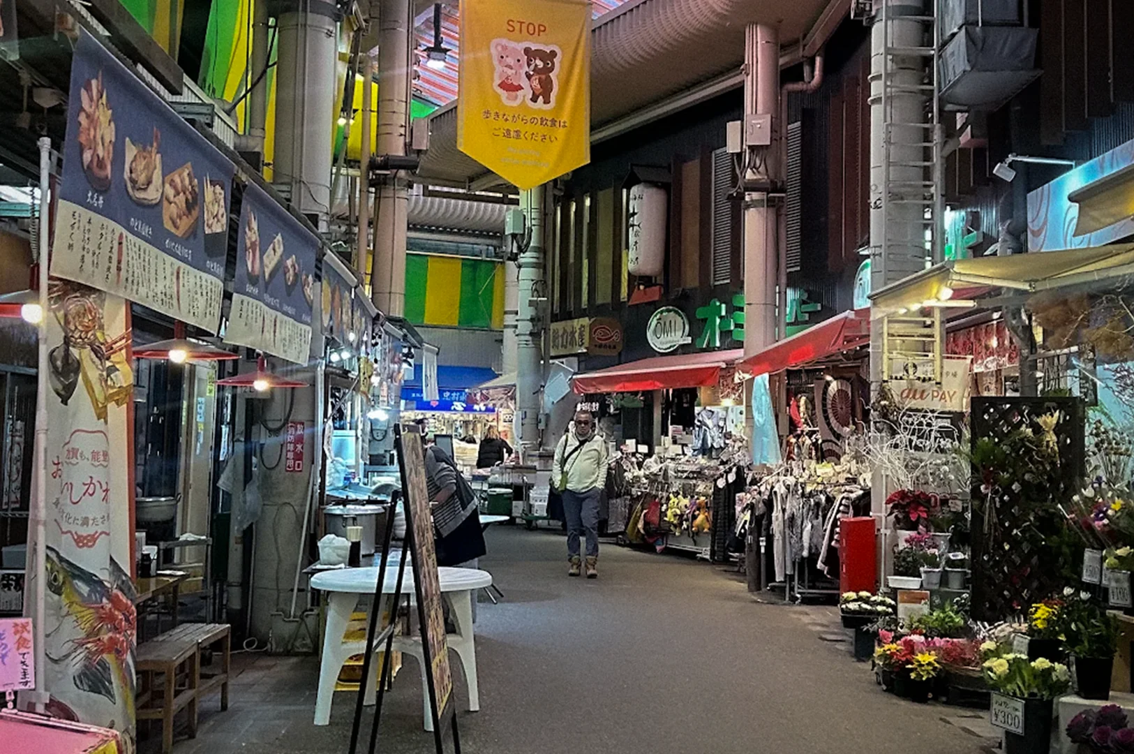 Covered shopping arcade at Omicho Market in Kanazawa, Japan, featuring seafood stalls, flower shops, local vendors, colorful storefronts, and a transparent roof canopy.