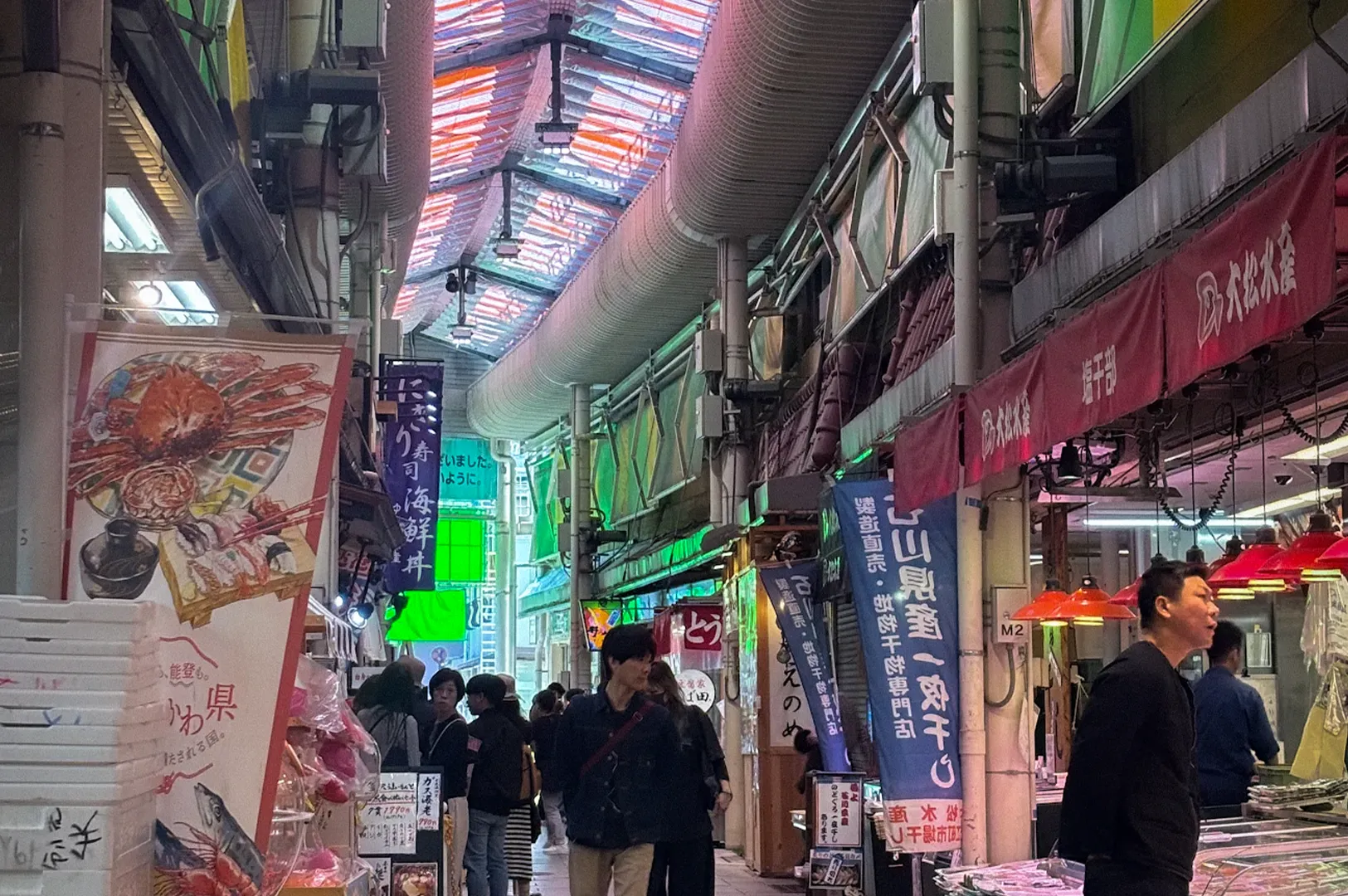 Interior of Omicho Market in Kanazawa, Japan, featuring seafood vendors, fresh fish displays, shoppers, and traditional market stalls beneath a colorful covered arcade roof.