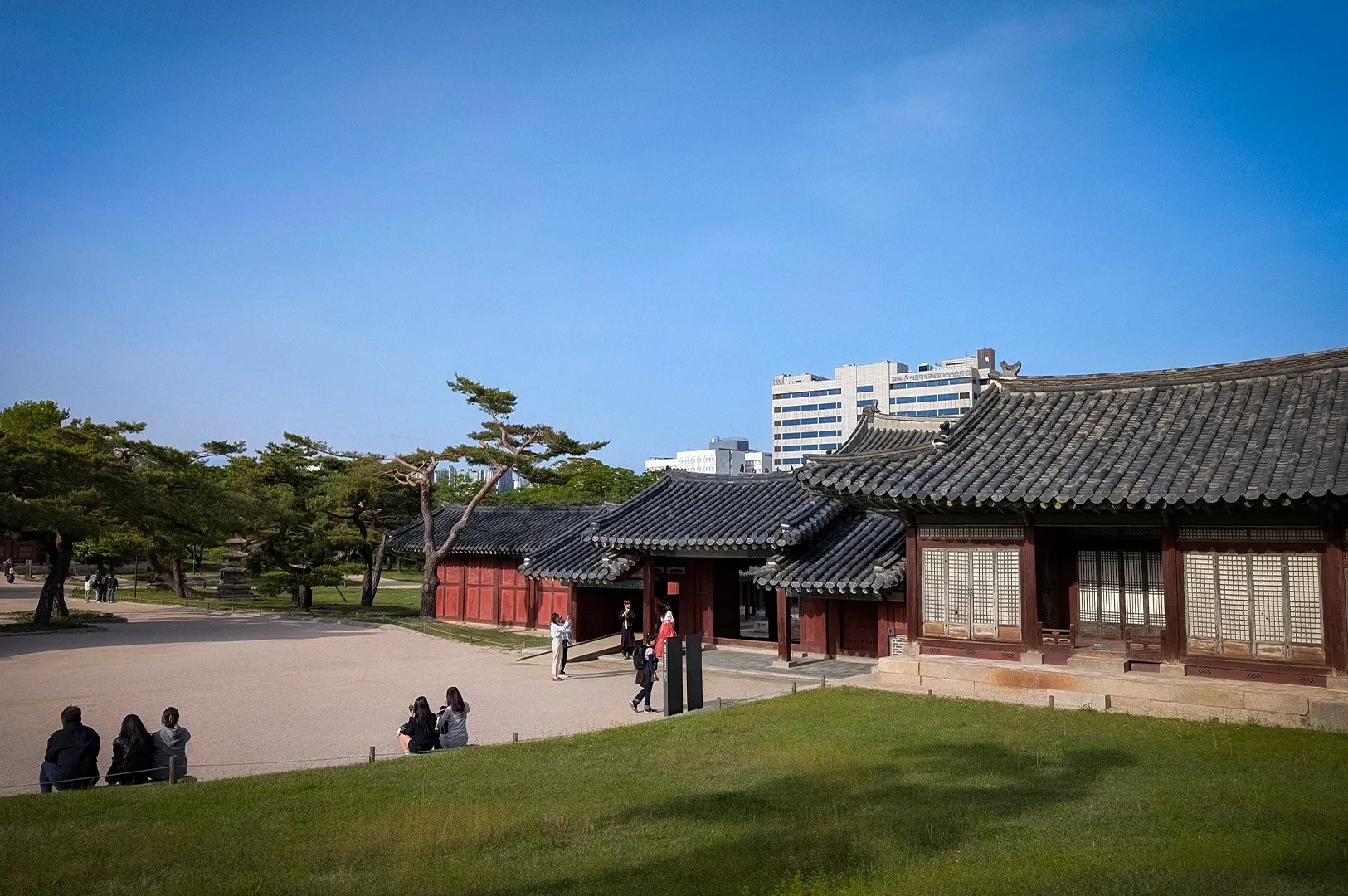 Traditional Korean palace courtyard featuring historic hanok-style buildings with tiled roofs, open grassy grounds, and visitors exploring the cultural heritage site against a backdrop of modern Seoul buildings and clear blue skies.
