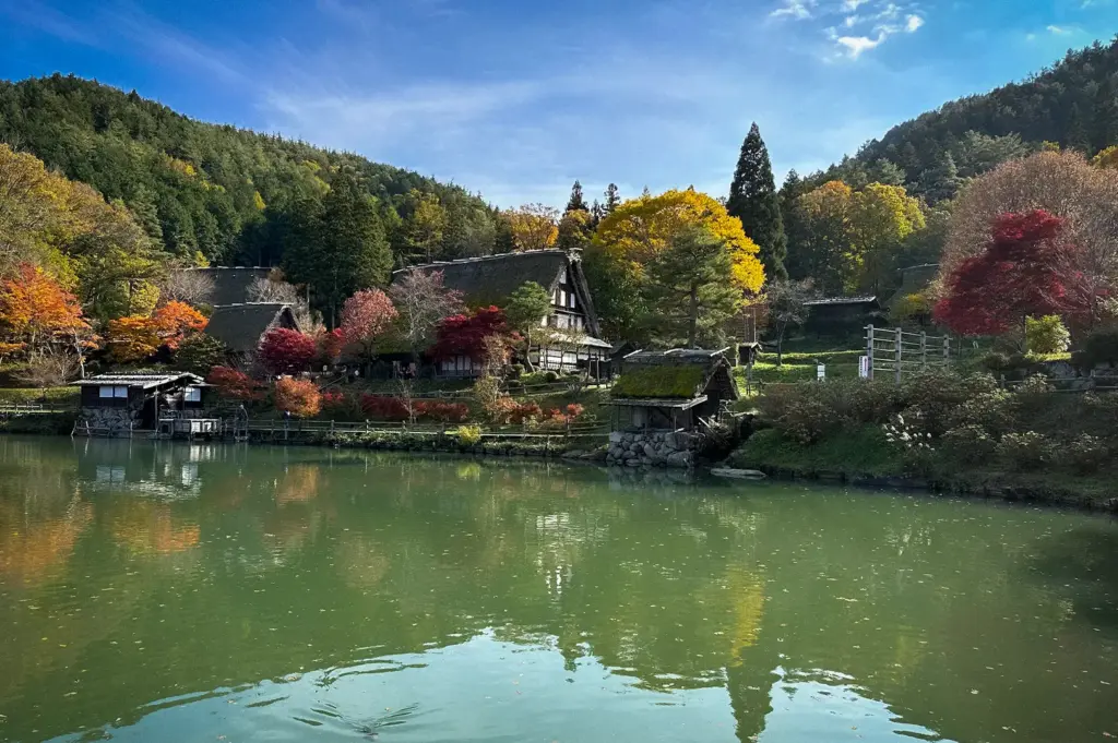 Hida no Sato Folk Village in Takayama, Japan, featuring traditional gassho-zukuri farmhouses, vibrant autumn foliage, and a tranquil pond surrounded by forested hills.