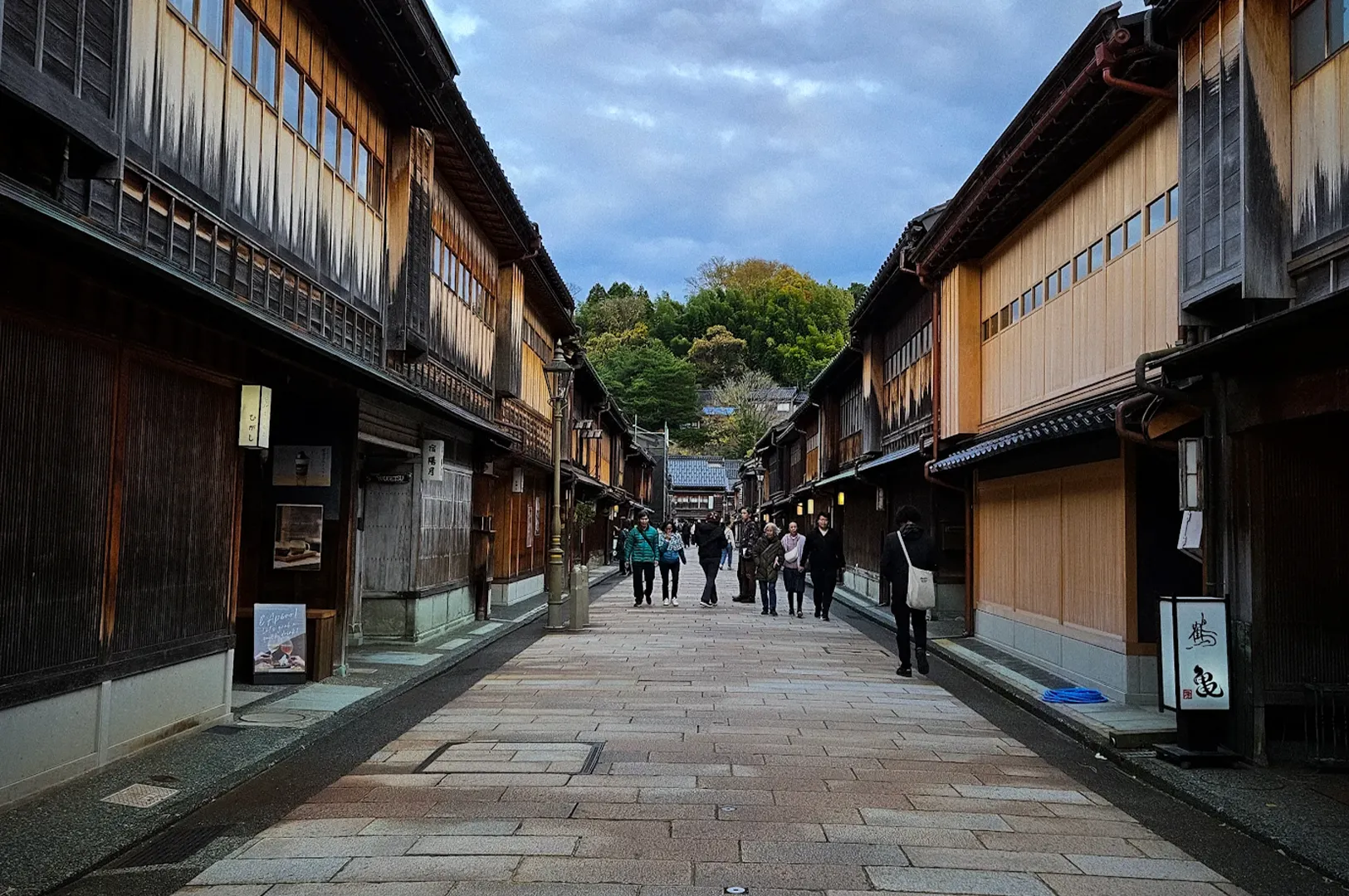 Traditional wooden teahouses lining the stone-paved street of Higashi Chaya District in Kanazawa, Japan, with visitors walking through the preserved historic geisha quarter under a cloudy sky.