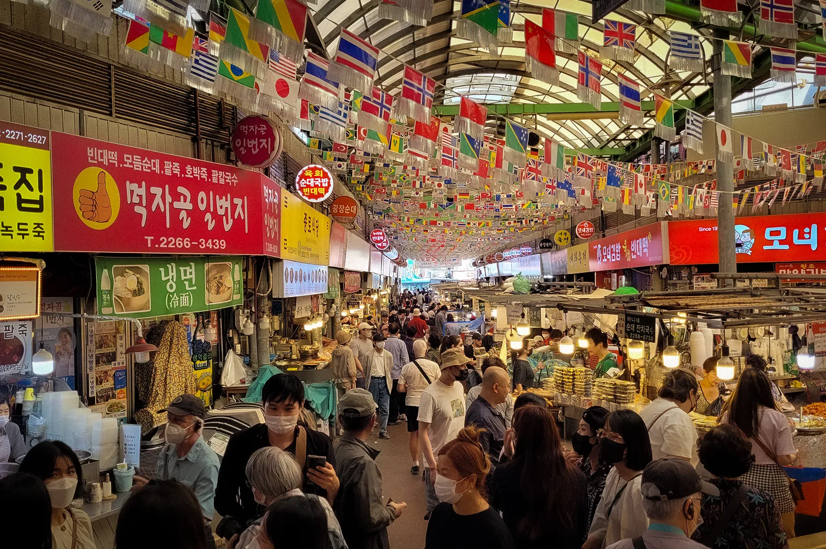 Crowded aisle inside Gwangjang Market in Seoul featuring traditional Korean food stalls, colorful international flags hanging overhead, illuminated shop signs, and visitors exploring one of South Korea’s most famous street food and shopping destinations.