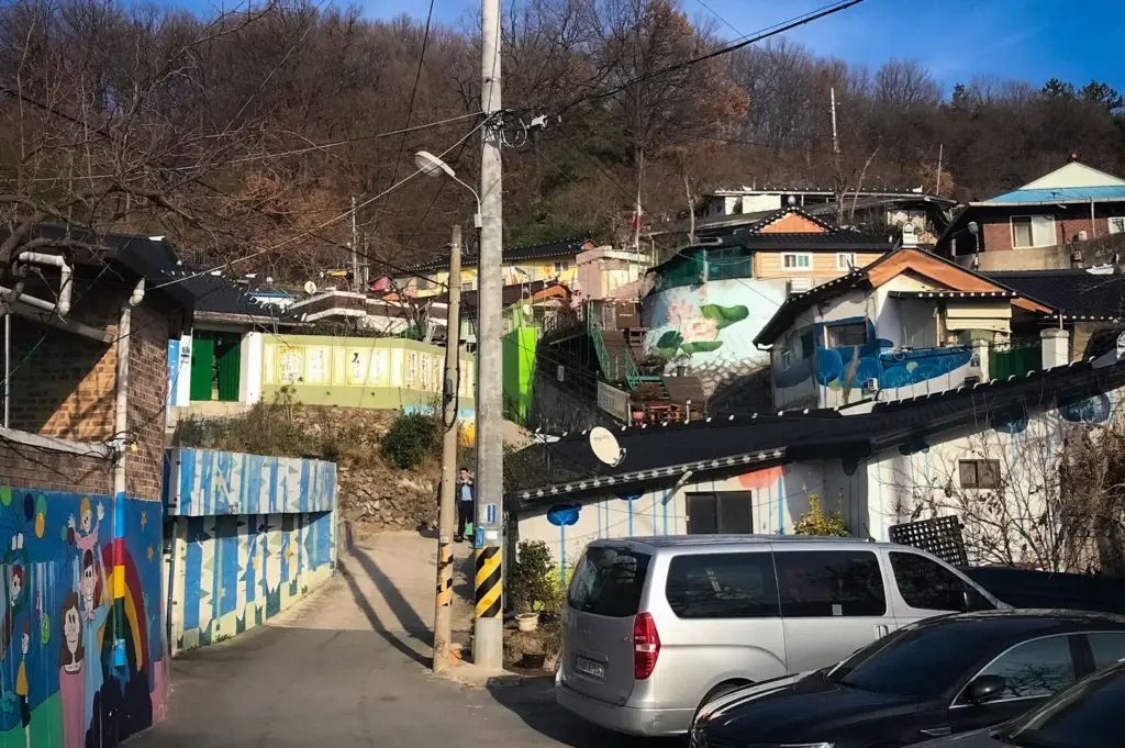 Quiet street in Jaman Mural Village, Jeonju, with colorful mural-painted houses, parked cars, and hillside homes under a clear blue sky.