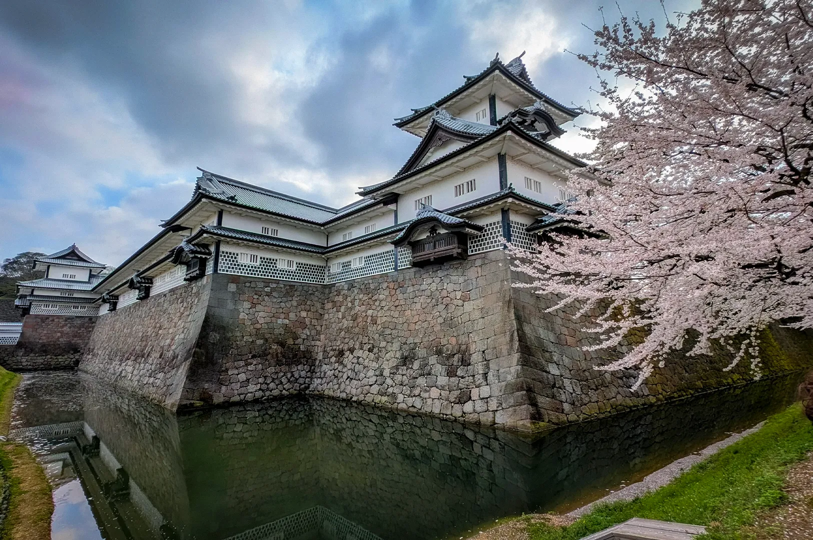 Historic Kanazawa Castle in Japan surrounded by blooming cherry blossom trees, with traditional white walls, stone fortifications, and a reflective moat under a cloudy spring sky.