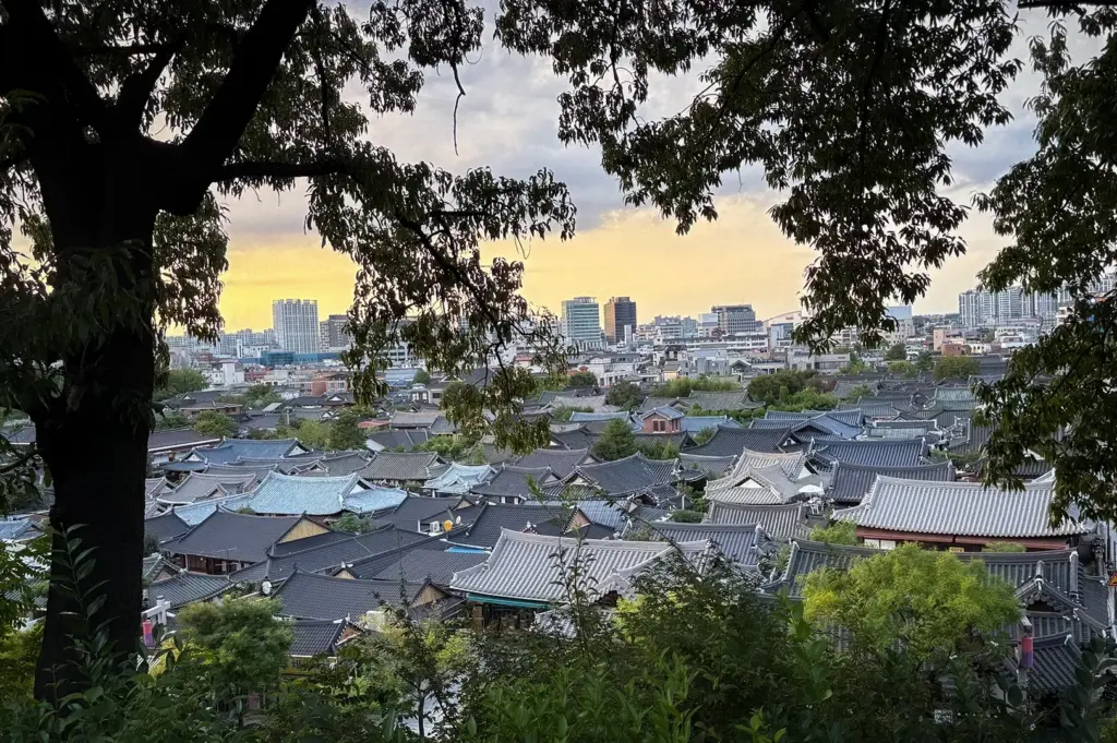Panoramic view of Jeonju Hanok Village with traditional Korean rooftops, colorful autumn foliage, modern city buildings, and forested mountains in the background.