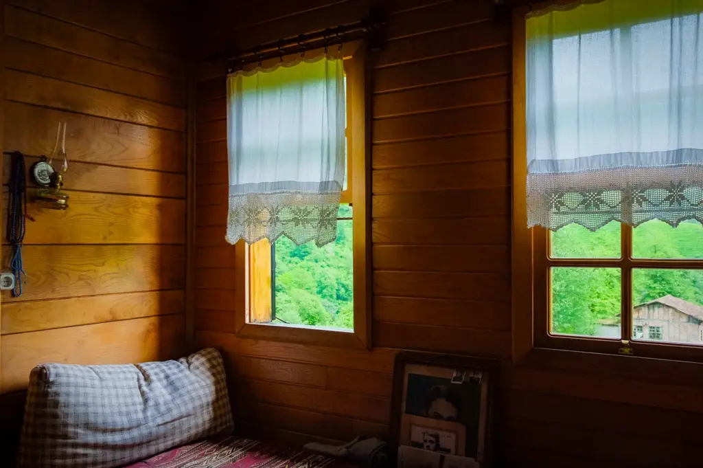 Cozy rustic wooden cabin bedroom with timber walls, plaid bedding, vintage oil lamp, lace curtains, and sunlit windows overlooking lush green countryside.