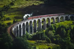 Steam train crossing the Glenfinnan Viaduct in the Scottish Highlands, surrounded by rolling green hills and scenic countryside.