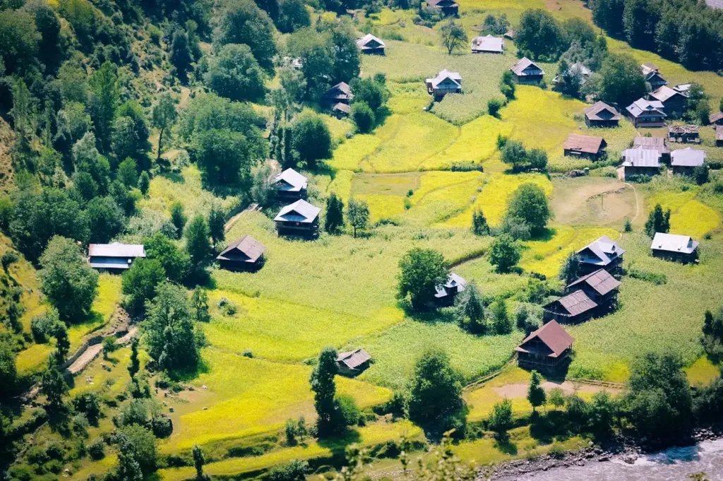 Aerial view of a rural village surrounded by lush green farmland, scattered homes, rolling fields, and dense trees beside a river in a scenic countryside landscape.
