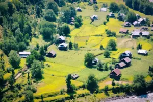 Aerial view of a rural village surrounded by lush green farmland, scattered homes, rolling fields, and dense trees beside a river in a scenic countryside landscape.