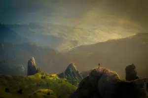 Panoramic mountain landscape with dramatic rock formations, rolling green hills, and a lone hiker standing on a rocky summit during golden hour.