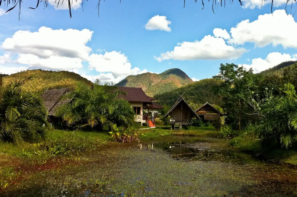 Scenic mountain village in Pai, Thailand featuring traditional houses, lush tropical greenery, a reflective pond, and rolling hills under a bright blue sky with white clouds.