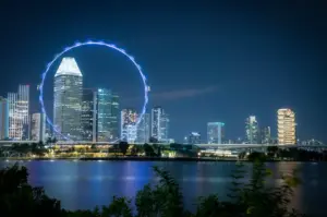 Night view of the Singapore Flyer and Marina Bay skyline with illuminated skyscrapers reflected on the waterfront under a clear evening sky.