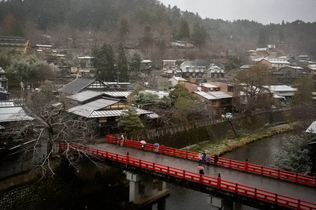 Scenic view of Takayama, Japan featuring the iconic red Nakabashi Bridge, traditional riverside buildings, and a light snowfall against forested hills in the historic old town.
