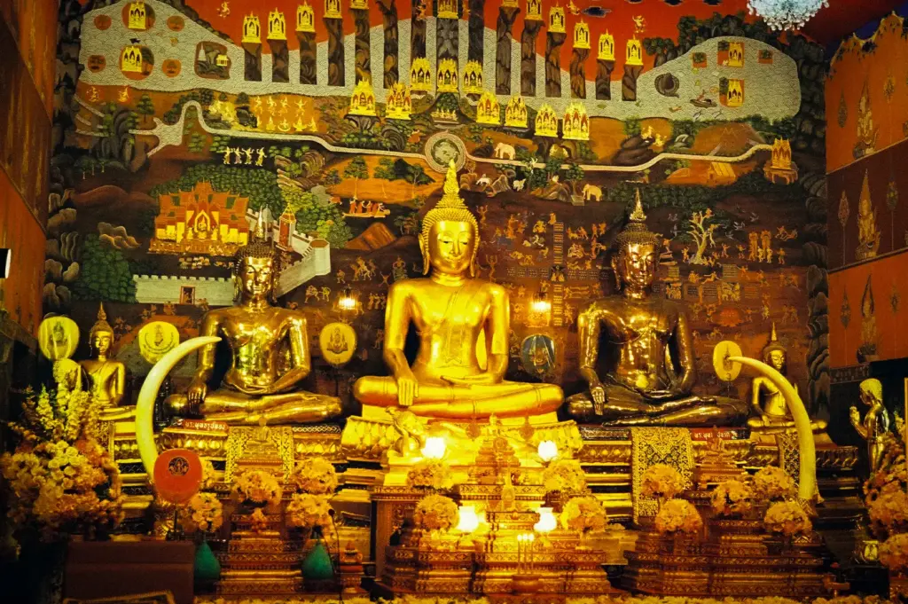 Golden Buddha statues inside Wat Phanan Choeng in Ayutthaya, Thailand, surrounded by ornate murals, floral offerings, and traditional Thai temple decorations.