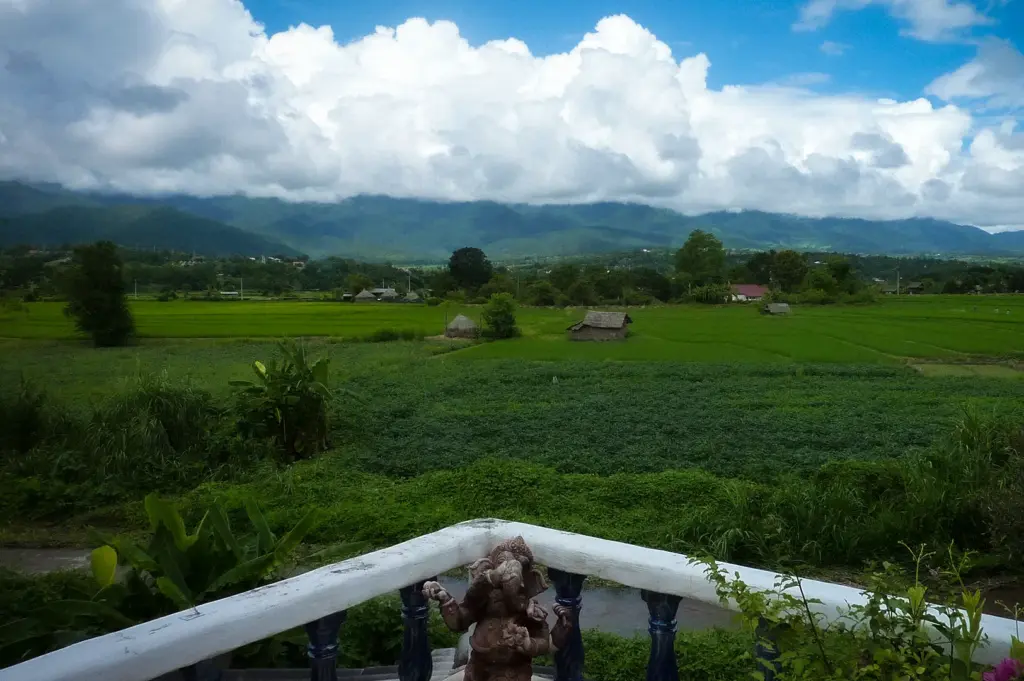 Panoramic view of Pai, Thailand showcasing lush green rice fields, distant mountain ranges, dramatic clouds, and a peaceful rural landscape viewed from a terrace.