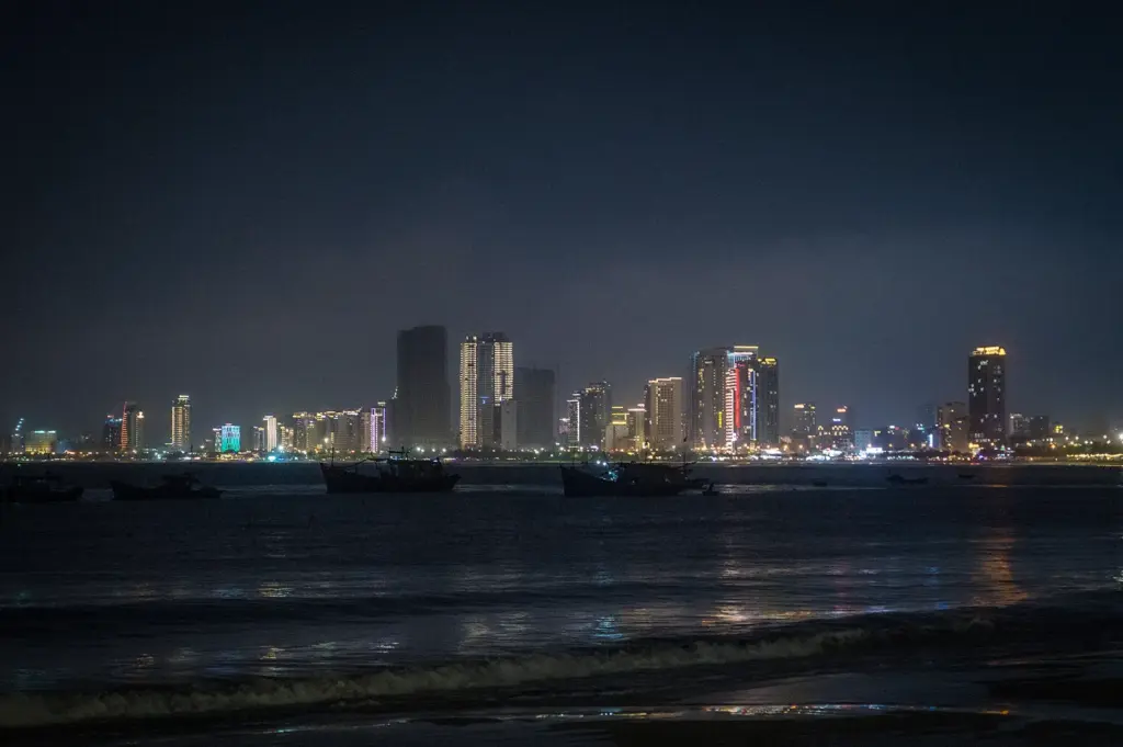 Night view of Da Nang, Vietnam skyline with illuminated high-rise buildings, fishing boats on the water, and reflections along the beachfront.