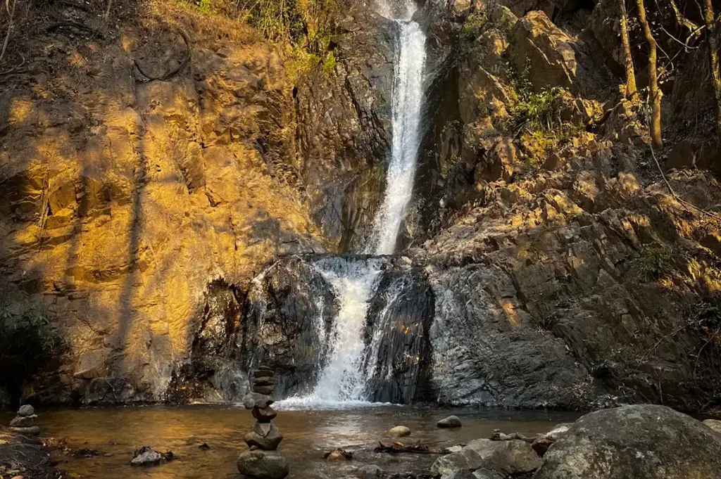 Mae Yen Waterfall in Pai, Thailand featuring a cascading waterfall flowing over rocky cliffs into a clear natural pool surrounded by forest and rugged boulders.