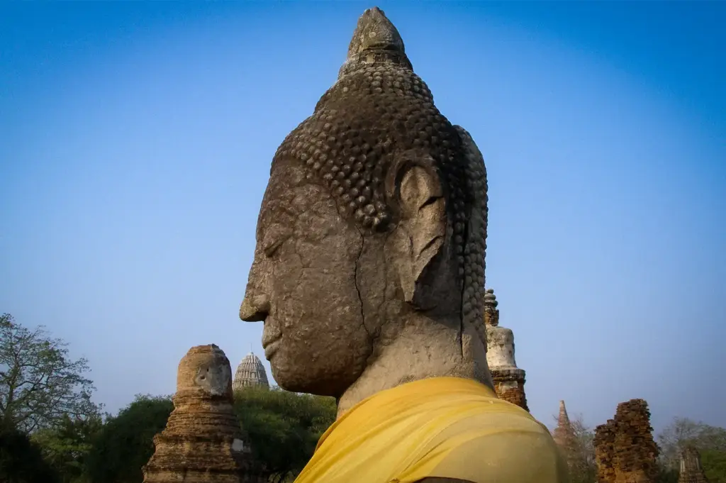 Ancient Buddha statue draped in a saffron robe at Ayutthaya Historical Park in Thailand, surrounded by centuries-old temple ruins and archaeological landmarks under a clear blue sky.