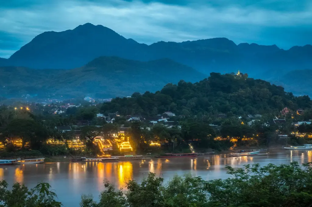 Evening view of Luang Prabang, Laos with the Mekong River, Mount Phou Si, lush mountains, and glowing riverside lights at sunset.