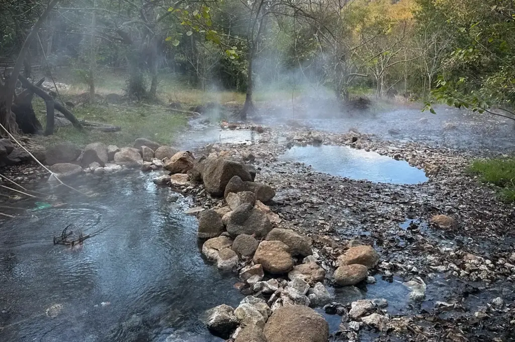 Tha Pai Hot Springs in Pai, Thailand with steaming geothermal pools, natural rock formations, forest surroundings, and clear blue sky creating a peaceful outdoor hot spring landscape.