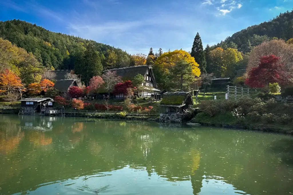 Scenic view of Hida no Sato Folk Village in Takayama, Japan, featuring traditional thatched-roof farmhouses, a tranquil pond, vibrant autumn foliage, and surrounding forested mountains.