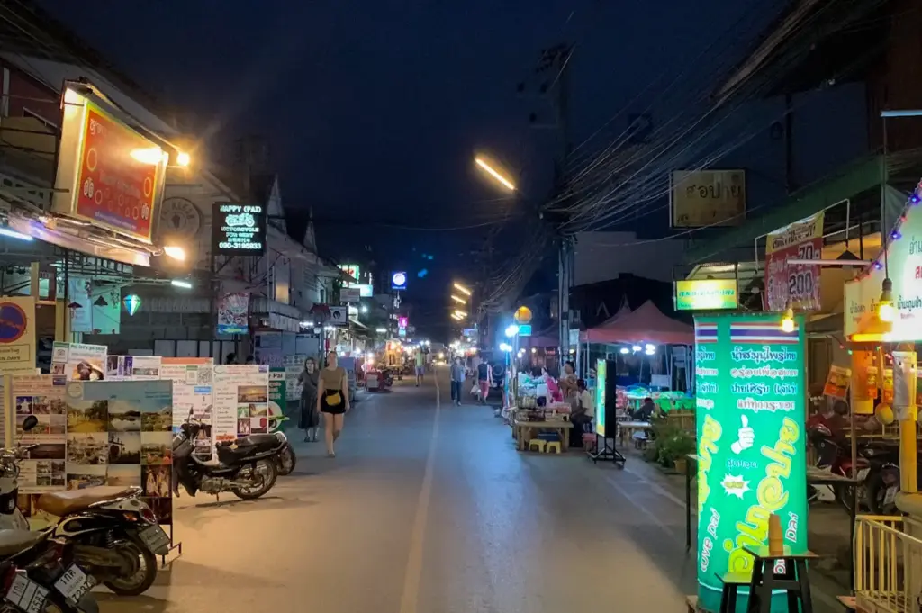 Pai Night Market in Pai, Thailand featuring a lively evening street lined with food stalls, local shops, colorful signs, motorbikes, and visitors exploring the vibrant night market atmosphere.