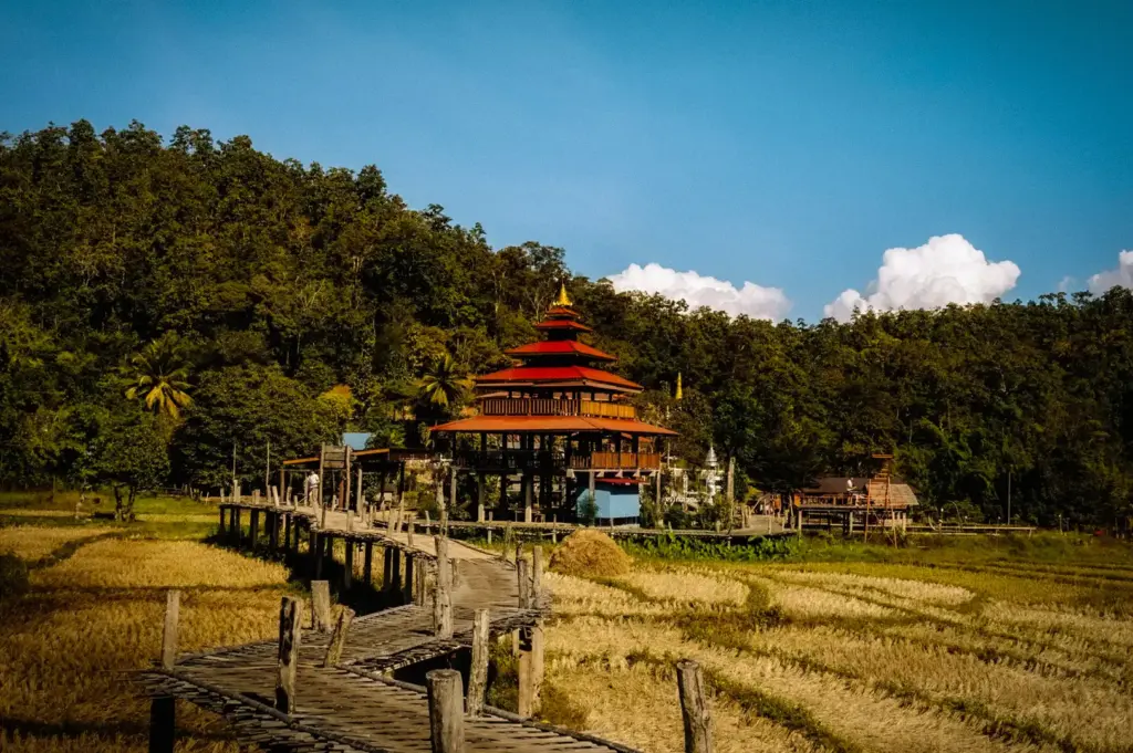 Traditional wooden bridge leading to a multi-tiered pavilion in Pai, Thailand, surrounded by harvested rice fields, lush forested hills, and a peaceful rural countryside landscape.