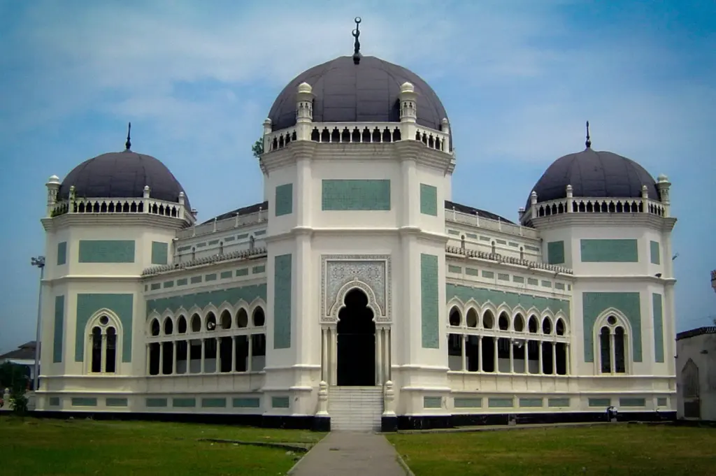 Front view of the Great Mosque of Medan in Indonesia showcasing its grand white facade, black domes, Islamic architecture, and landscaped grounds.