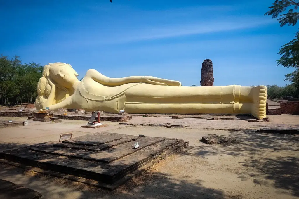 Reclining Buddha statue at Wat Lokayasutharam in Ayutthaya Historical Park, Thailand, with ancient temple ruins and a clear blue sky in the background.