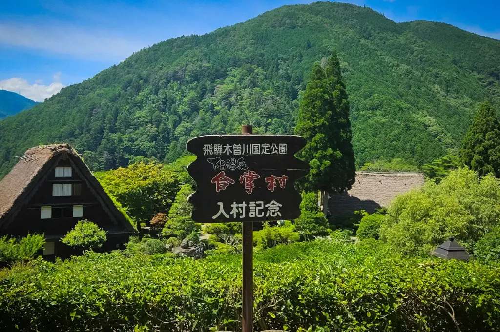 Scenic view near Gero Onsen featuring traditional thatched-roof houses, lush green mountains, and a village entrance sign in Japan's picturesque Hida region.