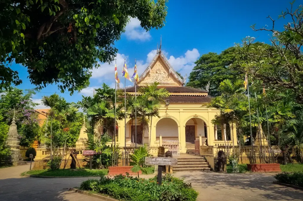 Front view of Wat Damnak temple in Siem Reap, Cambodia, surrounded by tropical gardens, palm trees, and traditional Khmer architecture under a clear blue sky.