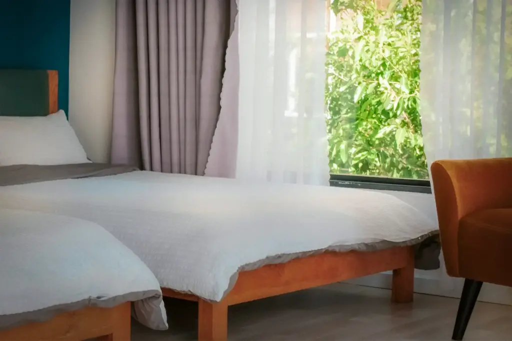 Bright modern homestay bedroom with twin beds, wooden bed frames, large window, sheer curtains, comfortable seating, and abundant natural daylight creating a relaxing stay.