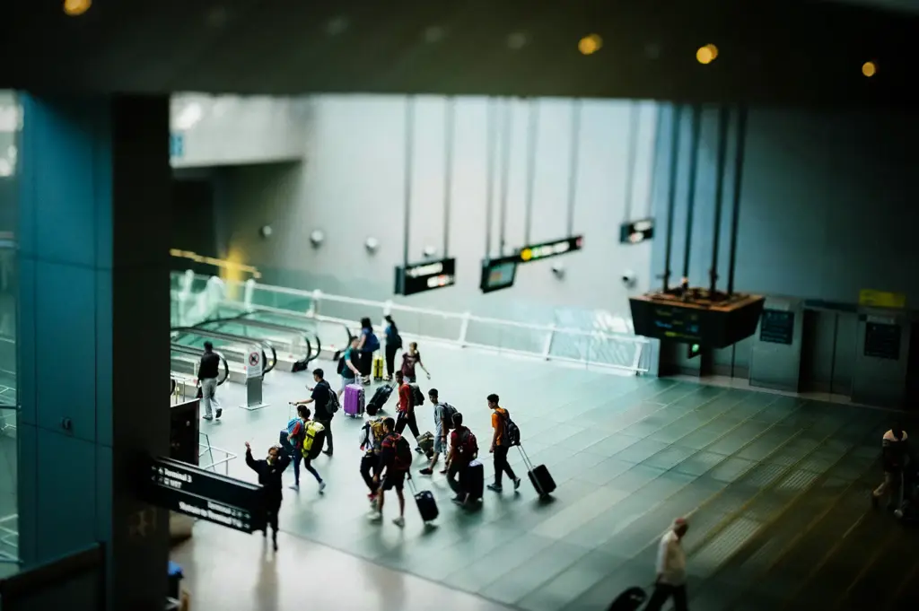 Busy airport terminal with travelers carrying luggage near escalators, directional signs, and modern departure facilities.